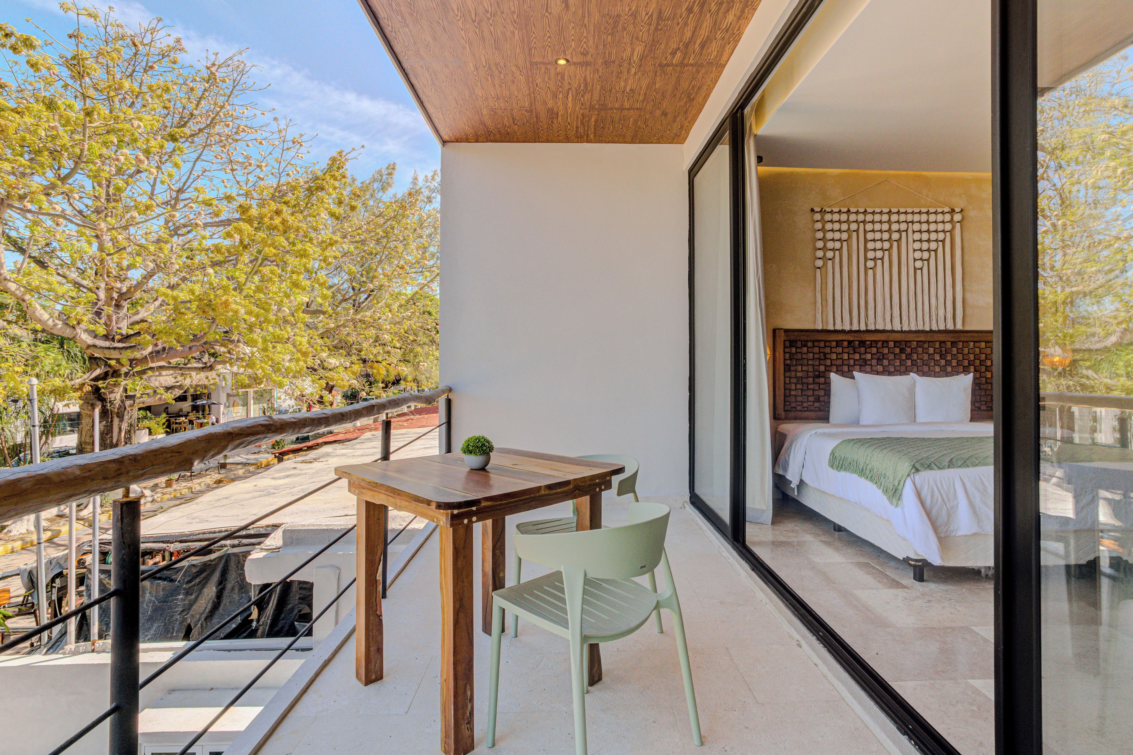 Un balcon avec une table et des chaises en bois, une vue sur un arbre et un aperçu d’une chambre à coucher avec un lit et une tête de lit.