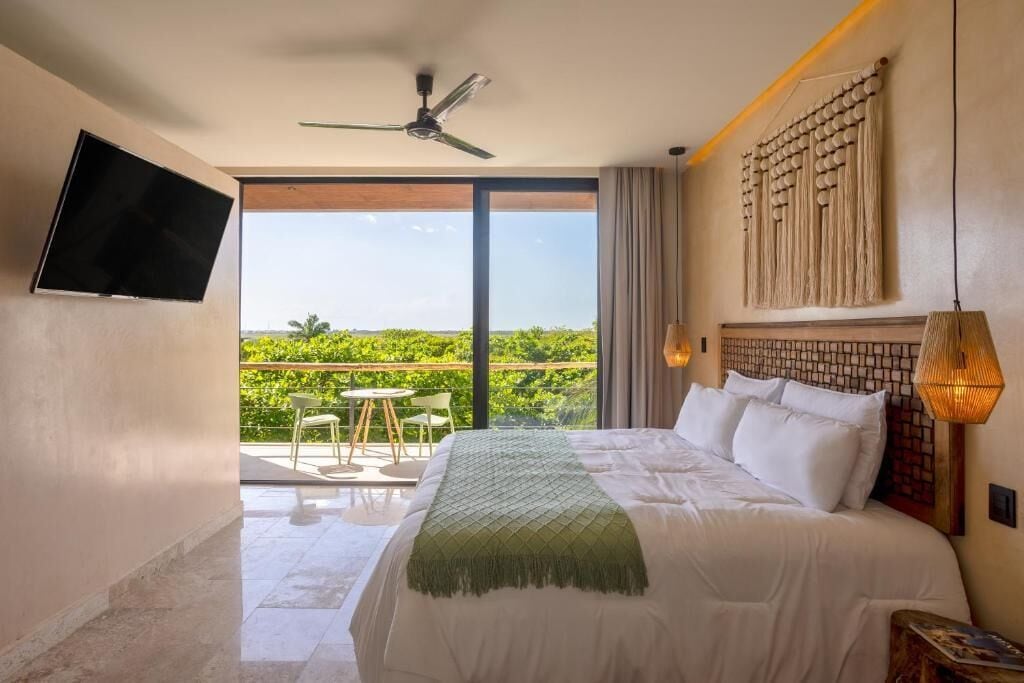 A hotel room with a bed, a television, a ceiling fan, a balcony with a view of greenery, and a decorative wall piece.