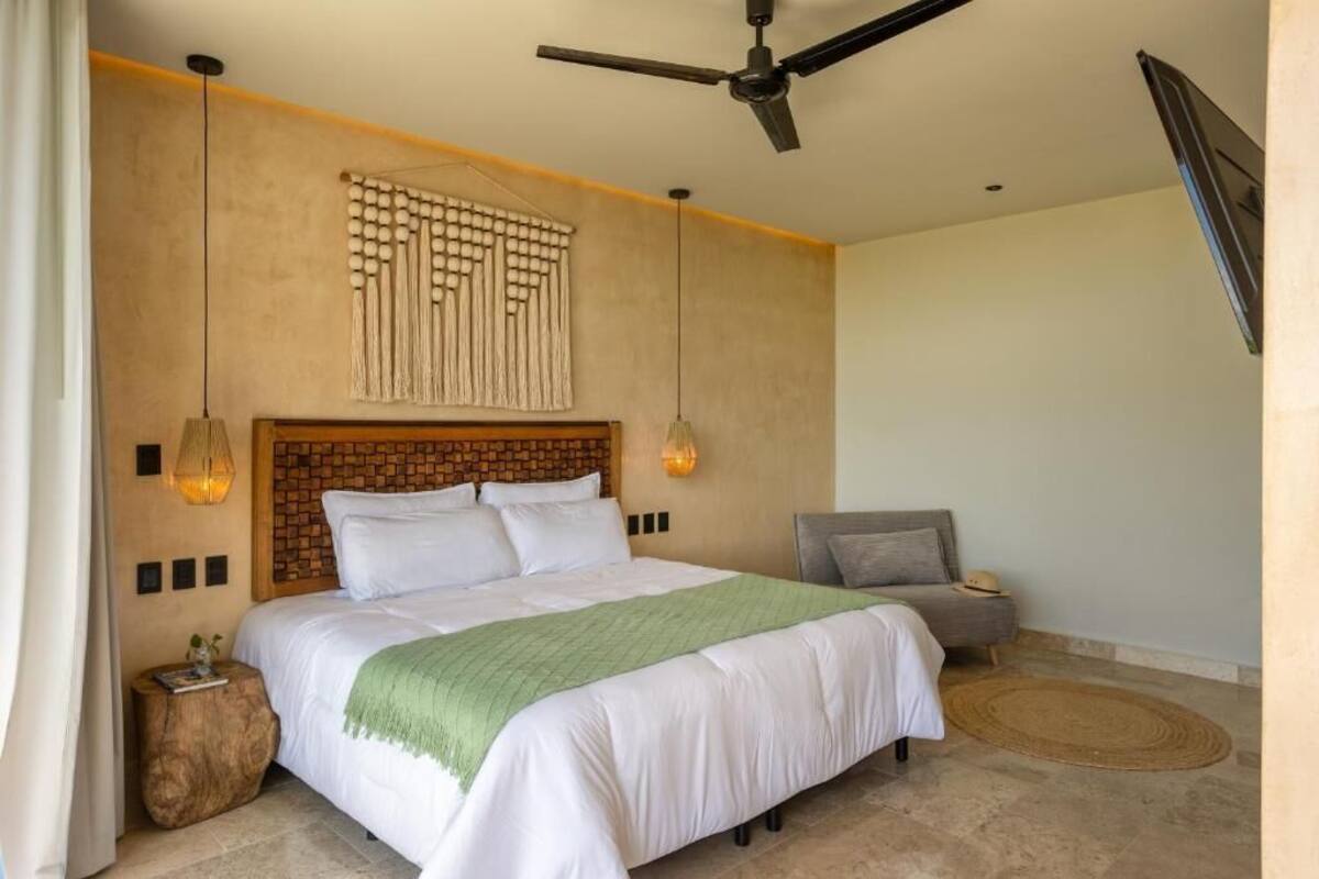 Quarto com cabeceira em padrão de tijolos, cama com roupa de cama branca, poltrona cinza, tapete redondo de fibras naturais e ventilador de teto.