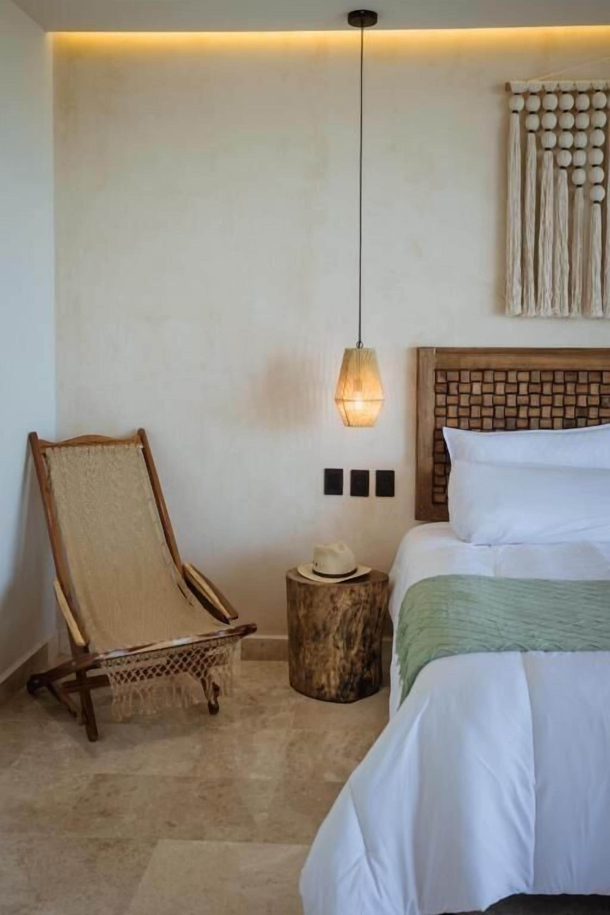 A bedroom with a bed, a chair, a wall-mounted light fixture, and a small round table with a hat.