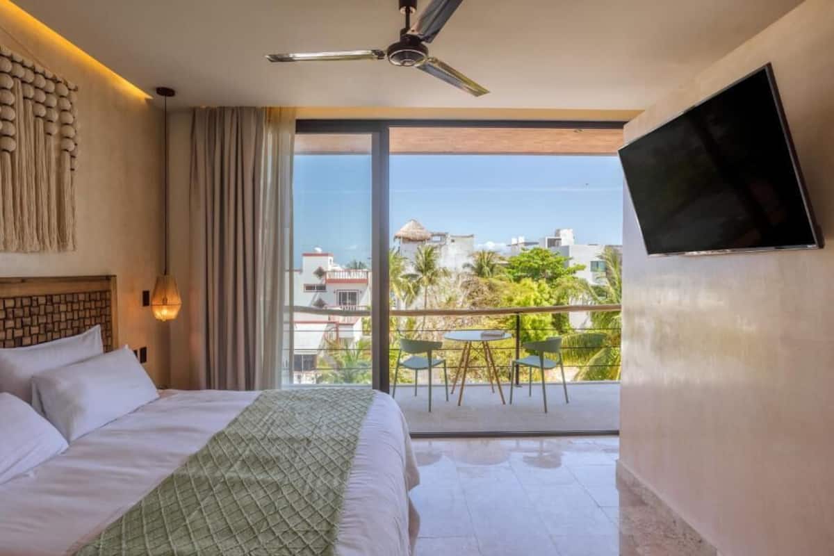 Quarto de hotel com cama, ventilador de teto, televisão, varanda com mesa e cadeiras, e vista para edifícios e árvores.