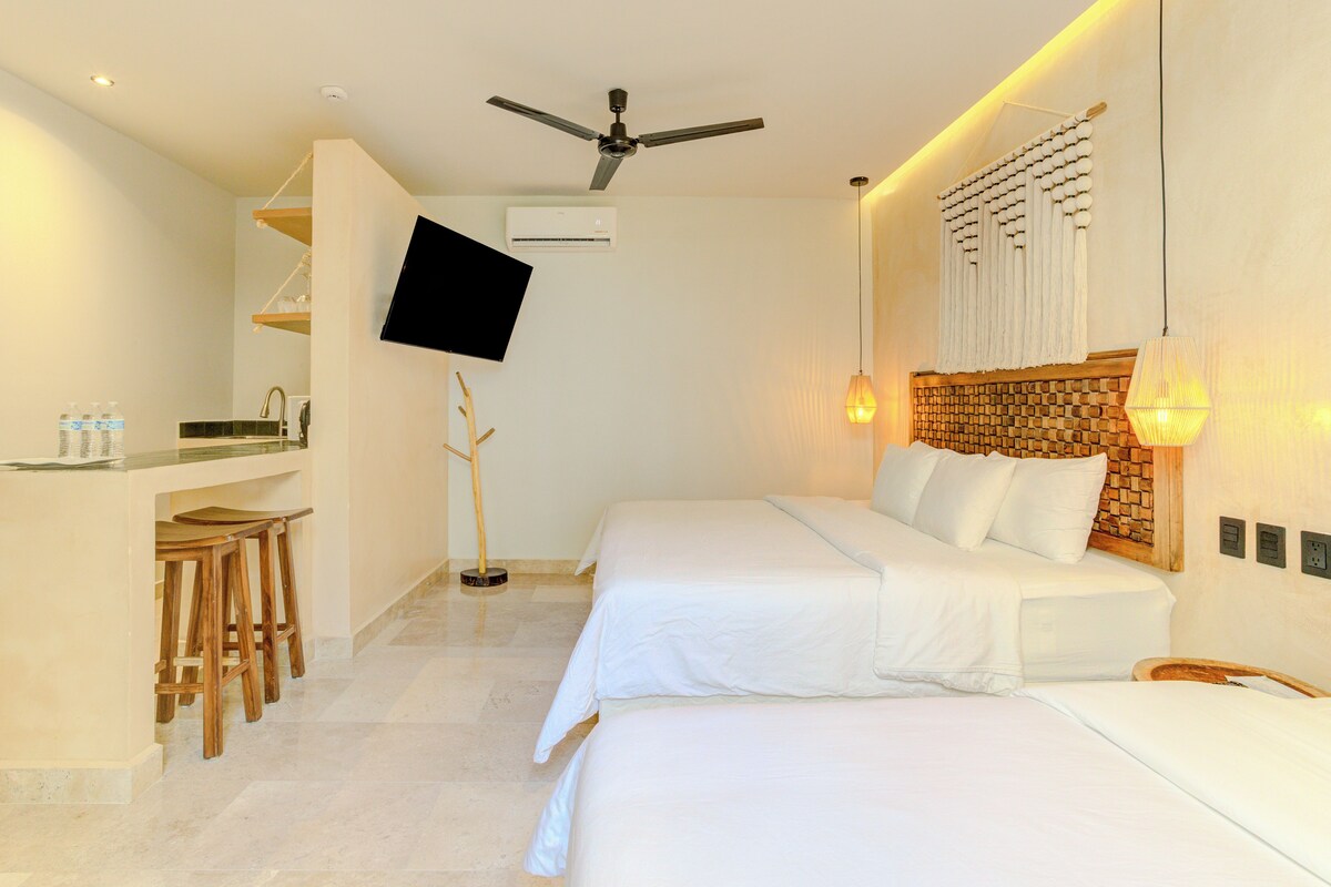 A hotel room with two beds, a TV mounted on the wall, a ceiling fan, and a small kitchenette area.