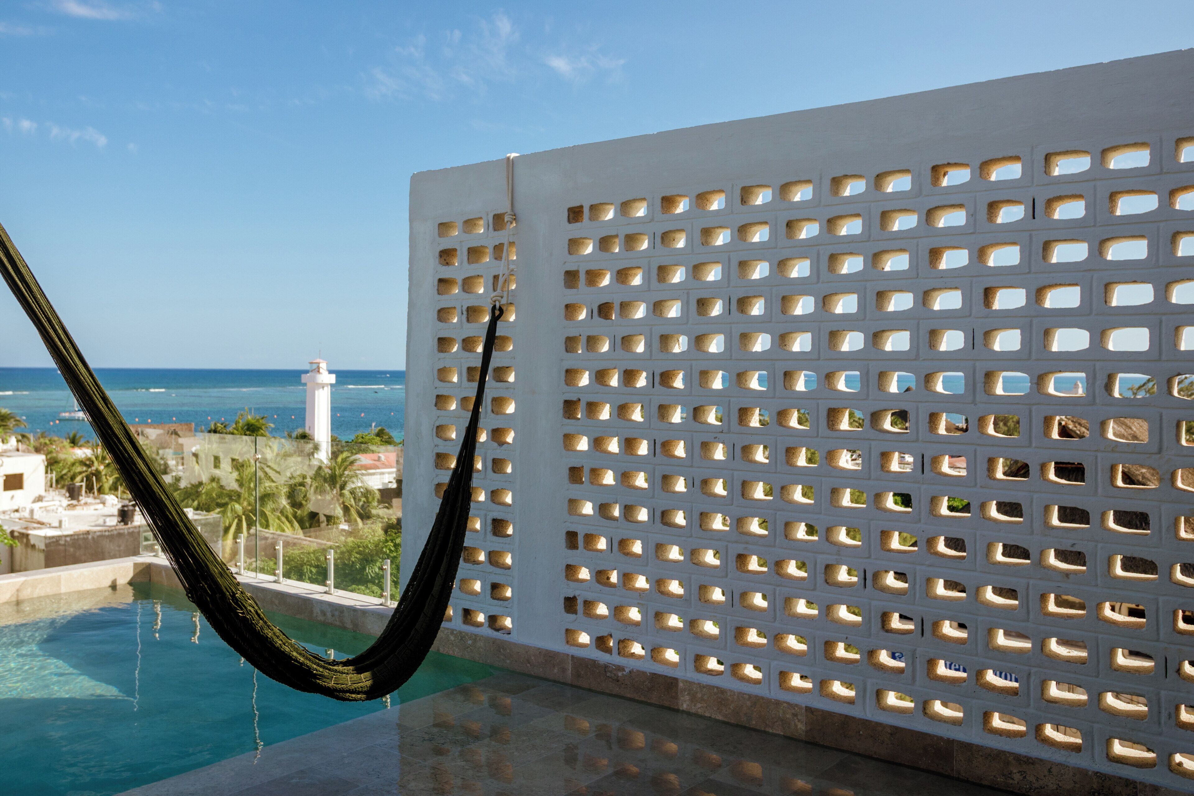 A pool with a hammock, a white perforated wall, and a view of the ocean and buildings.