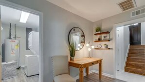 Una habitación con una mesa de madera, una silla, un espejo y una estantería con plantas y un cartel.
