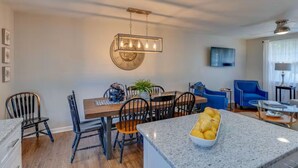 Una cocina moderna con encimeras de granito, un comedor con mesa y sillas de madera, y una sala de estar con sillones azules y un televisor de pantalla plana.
