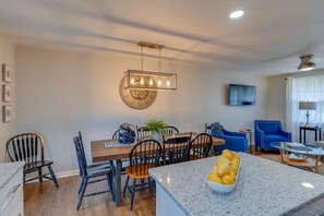 Eine moderne Küche mit Granit-Arbeitsplatten, ein Essbereich mit Holztisch und -stühlen sowie ein Wohnzimmer mit blauen Sesseln und einem Flachbildfernseher.