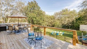 Una terraza de madera con mobiliario exterior, una parrilla y vistas a una zona verde con árboles.