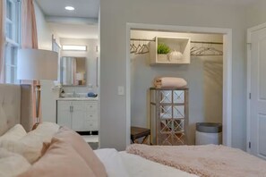 Ein Schlafzimmer mit einem Bett, einer Nachttischlampe, einem Schminktisch, einem Spiegel, einem Kleiderschrank mit Kleidung und einem kleinen Tisch mit einer Pflanze.