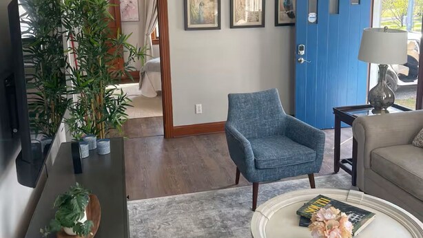 A living room with a blue door, a grey patterned rug, a white coffee table with flowers, a blue armchair, and a television on a cabinet. - The Haven near the lake and Nash! 3bd/2ba (Hendersonville)