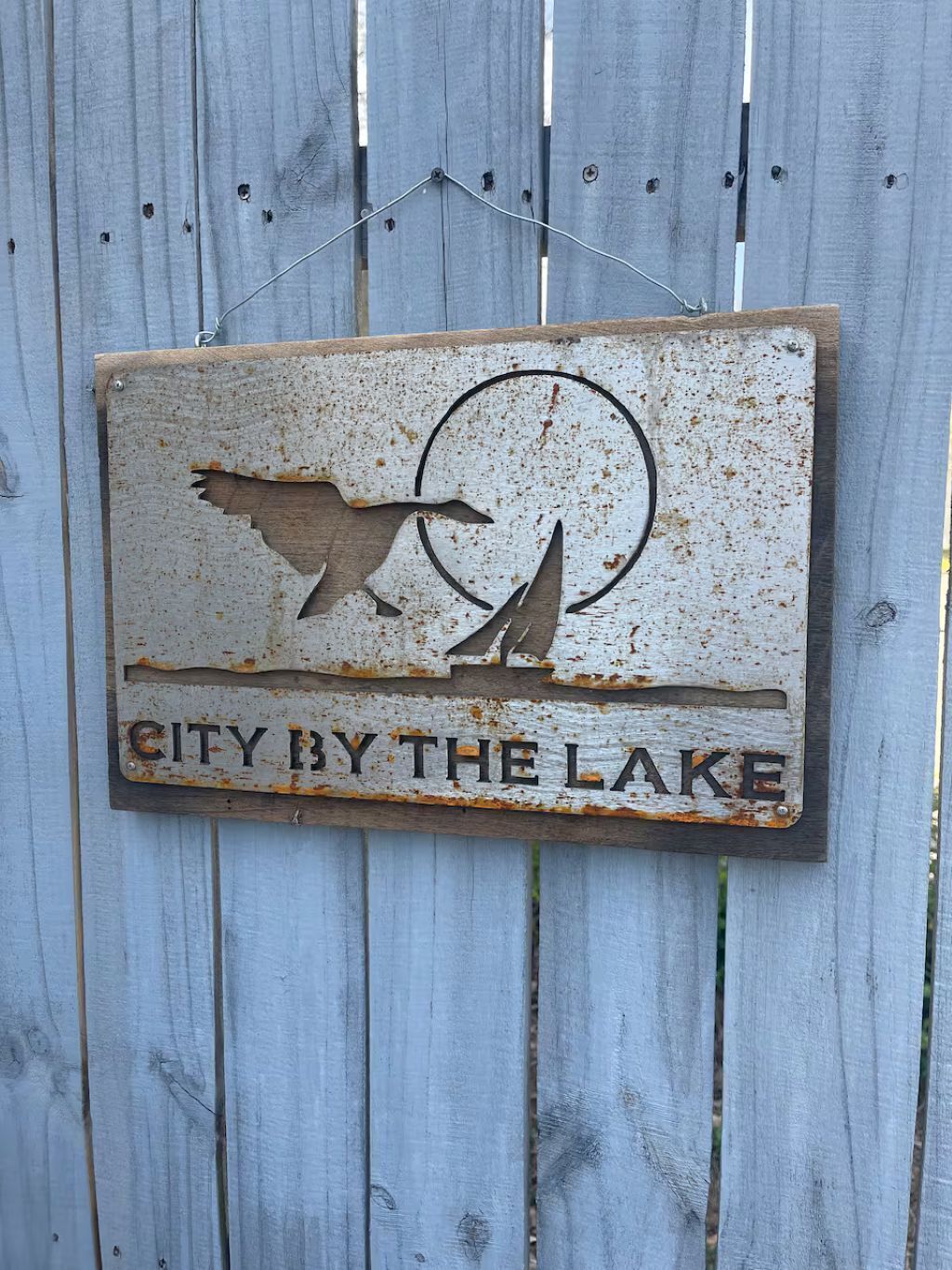 Uma placa de metal rústica com um pássaro e um barco, exibindo o texto 'CITY BY THE LAKE' sobre um fundo de madeira.