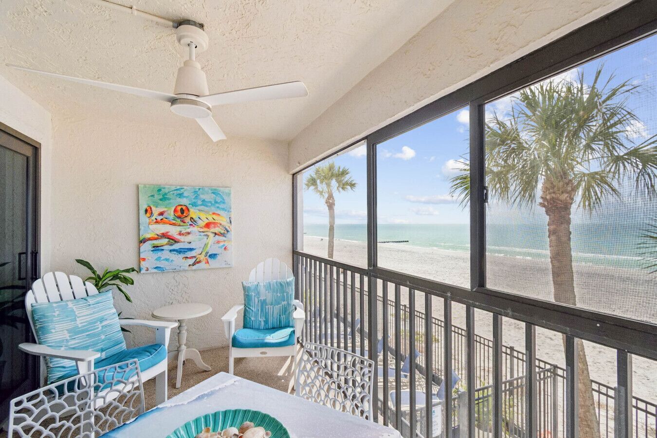 A balcony with a view of the beach, a ceiling fan, and a painting.