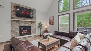 A living room with a stone fireplace, leather sofas, a wooden coffee table, and a large TV screen displaying 'NETFLIX'. - The Ultimate Social Stay - 6BRs, Hot Tub, Arcade, Fire Pit. SUMMER SAVINGS! (Albrightsville)