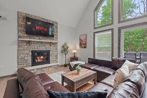 A living room with a stone fireplace, leather sofas, a wooden coffee table, and a large TV screen displaying 'NETFLIX'.