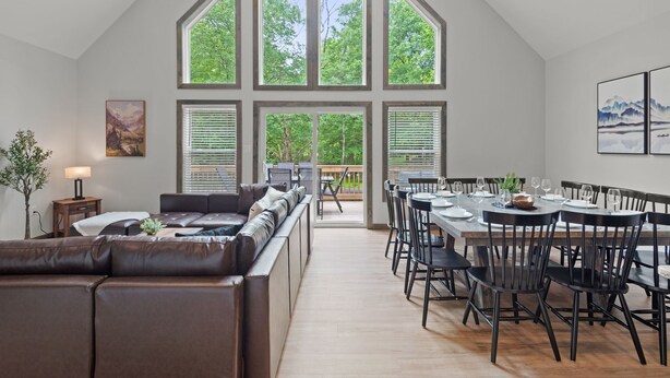 A spacious living area with a large window, a dining table with chairs, and a leather sofa. - The Ultimate Social Stay - 6BRs, Hot Tub, Arcade, Fire Pit. SUMMER SAVINGS! (Albrightsville)