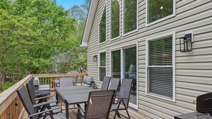 A deck with a table and chairs, a grill, and a house with large windows and a deck railing. - The Ultimate Social Stay - 6BRs, Hot Tub, Arcade, Fire Pit. SUMMER SAVINGS! (Albrightsville)
