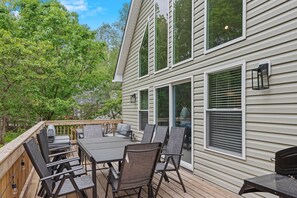 A deck with a table and chairs, a grill, and a house with large windows and a deck railing.
