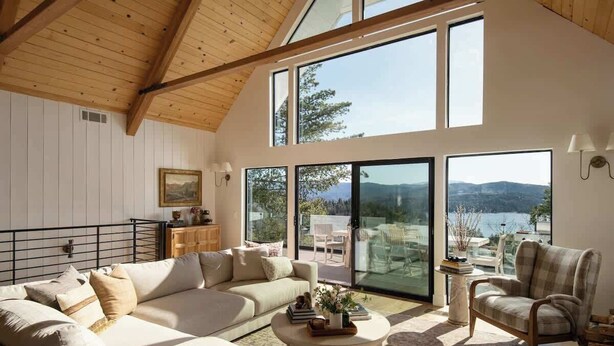 A spacious living room with a large sofa, a coffee table, and a chair. There are large windows offering a view of the outdoors. - Highland Haus (stunning lakeview + private dock!) (Lake Arrowhead)