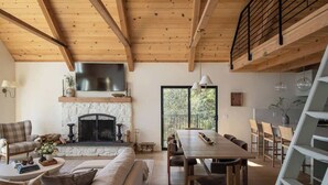A cozy living room with a fireplace, wooden beams, a dining area, and a view of the outdoors. - Highland Haus (stunning lakeview + private dock!) (Lake Arrowhead)