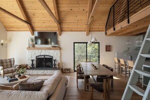 A cozy living room with a fireplace, wooden beams, a dining area, and a view of the outdoors. - Highland Haus (stunning lakeview + private dock!) (Lake Arrowhead)