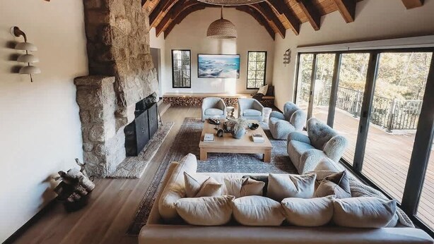 Un salon chaleureux avec une cheminée en pierre, des poutres apparentes en bois au plafond, une grande fenêtre et un coin salon confortable.