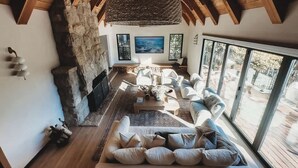 Un vaste salon doté d’une cheminée en pierre, de poutres apparentes en bois et de grandes fenêtres offrant une vue sur l’extérieur.