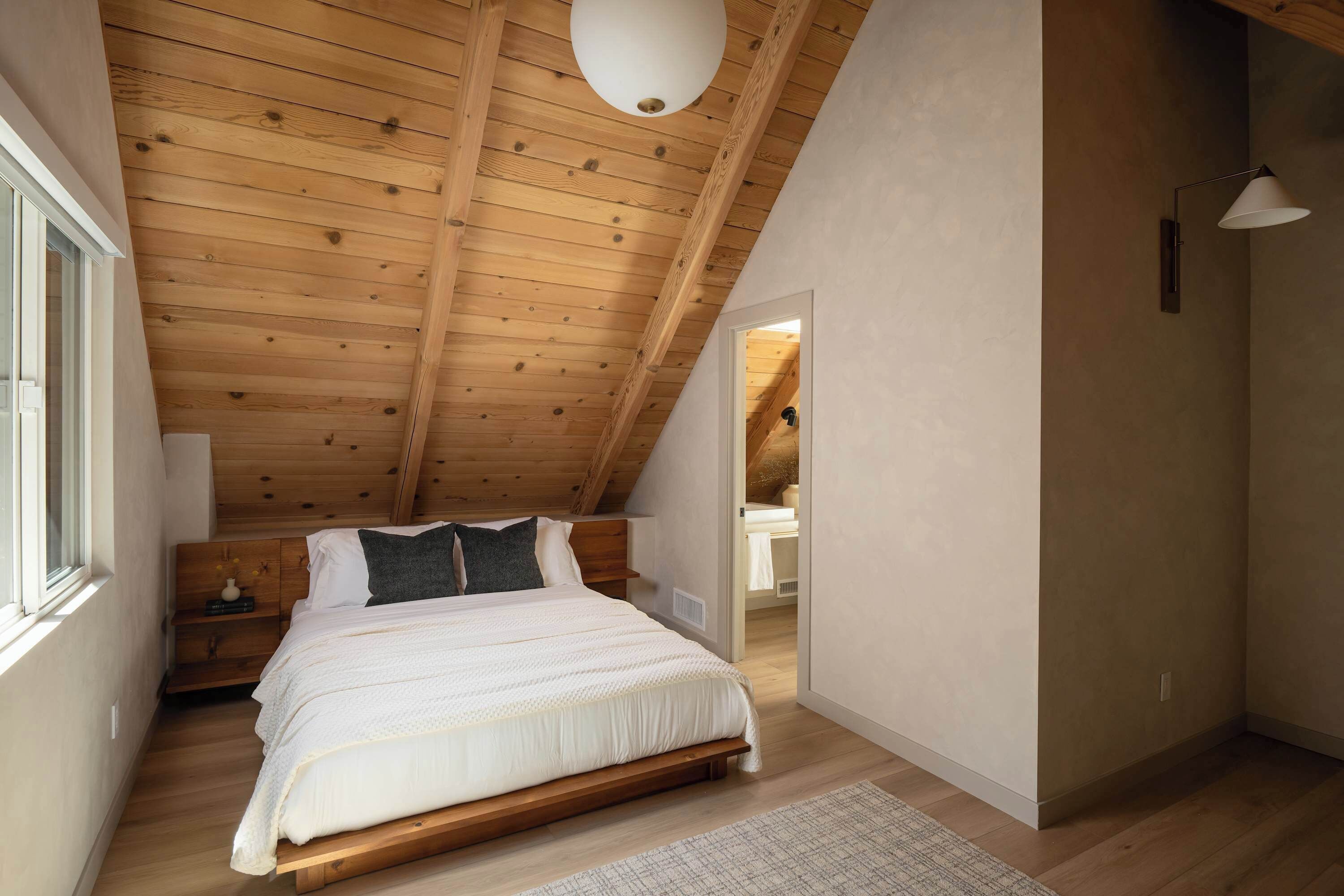 Quarto com teto inclinado, vigas de madeira, cama com roupa de cama branca, mesa de cabeceira e uma luminária.