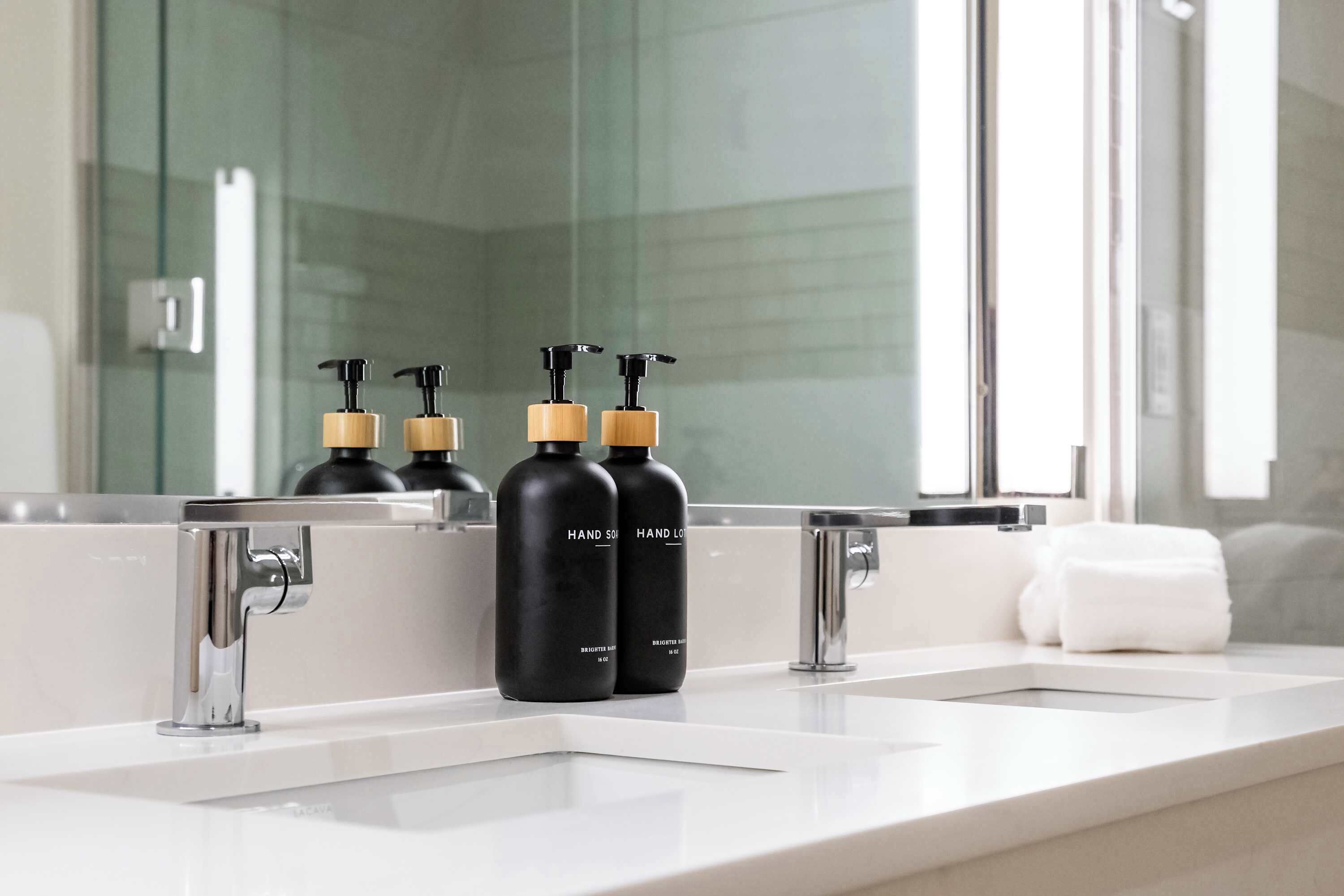 A bathroom with two sinks, a mirror, and three black bottles with pumps.