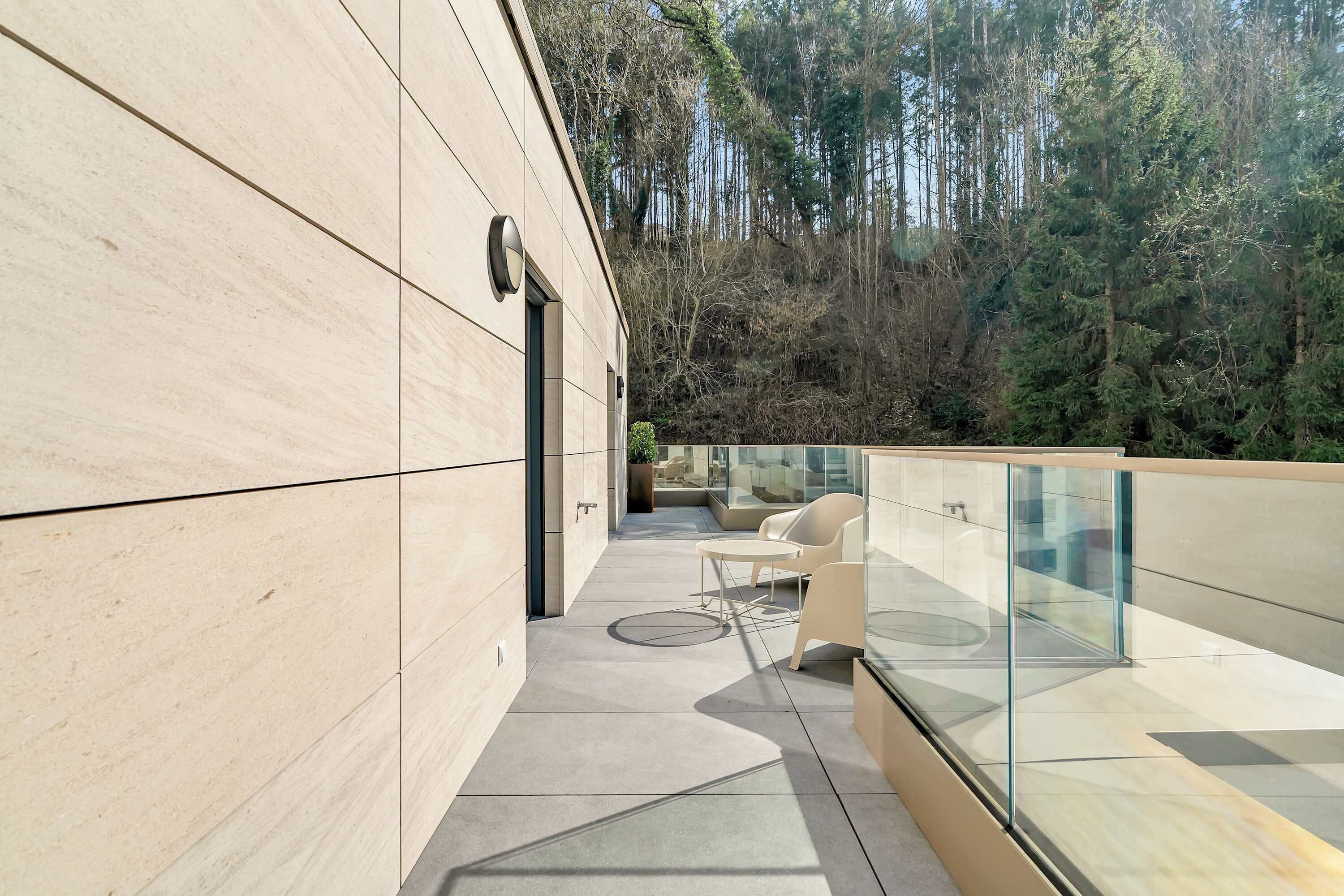 A modern building with a glass railing, a patio with chairs, and a view of a forested hillside.