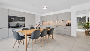 Een moderne keuken met ingebouwde apparatuur, een eethoek met een houten tafel en stoelen, en uitzicht door een glazen deur.