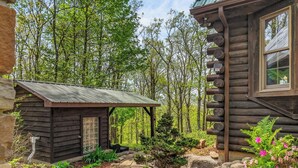 Une cabane rustique en rondins de bois, avec un petit abri de jardin, entourée d’un sentier pavé de pierres et d’une végétation luxuriante.