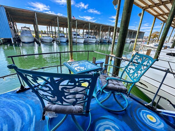 Un puerto deportivo con barcos amarrados, una zona de estar techada con sillas y una mesa, y un cielo despejado.