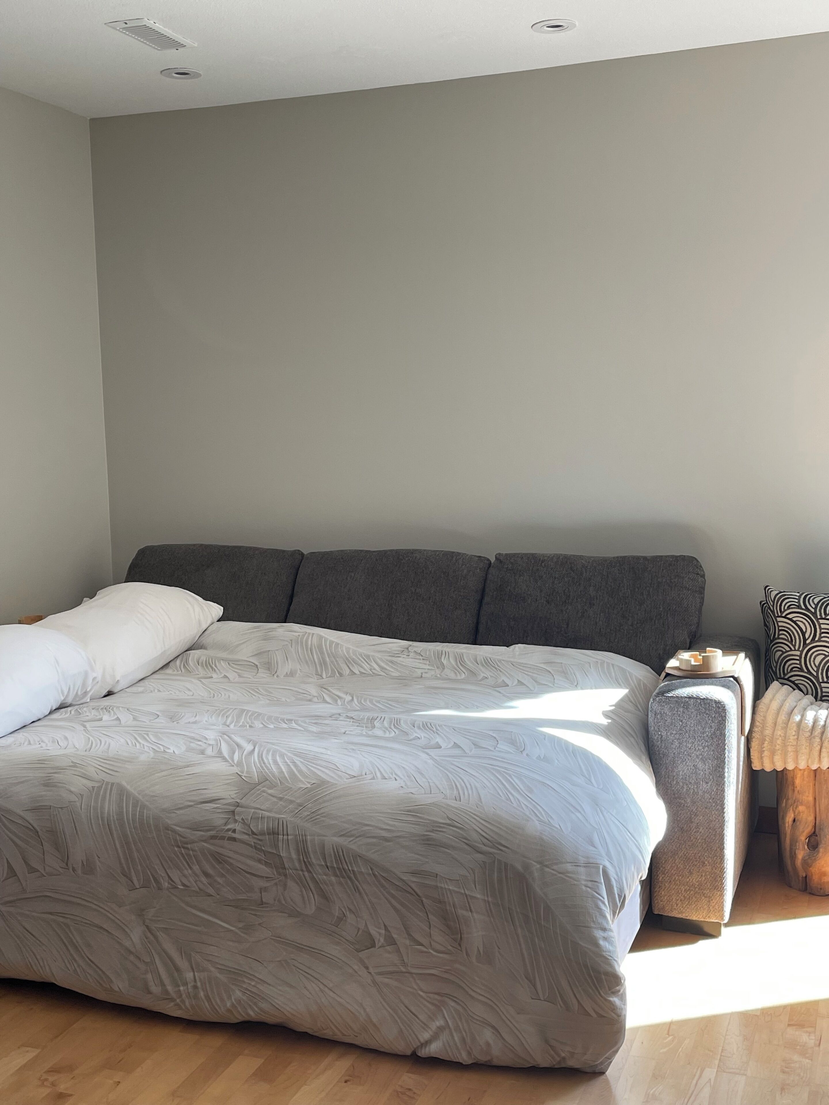 A large bed with a textured white cover, a grey headboard, and a wooden nightstand with a patterned pillow.
