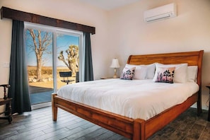 A bedroom with a large bed, a wooden headboard, a nightstand, a lamp, and a view of a desert landscape through a sliding glass door. - Pipes Canyon Retreat · 2 Homes, 10 Acres, Hot Tubs (Yucca Valley)