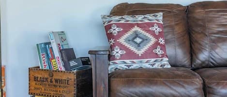 Una sala de estar acogedora con un sofá de cuero marrón, un cojín estampado y un caja de madera con libros y una botella.
