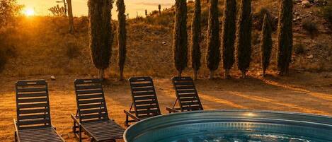 A hot tub surrounded by lounge chairs, with cypress trees and a sunset in the background.
