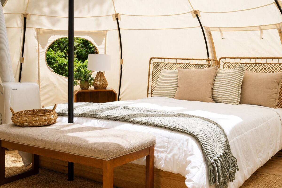 A tent-style room with a bed, a bench, a lamp, a basket, and a view of greenery outside.