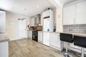 Eine moderne Küche mit weißen Schränken, einer Edelstahl-Dunstabzugshaube, einem Einbauofen und einem Kühlschrank. Zwei schwarze Barhocker stehen an der Arbeitsplatte.