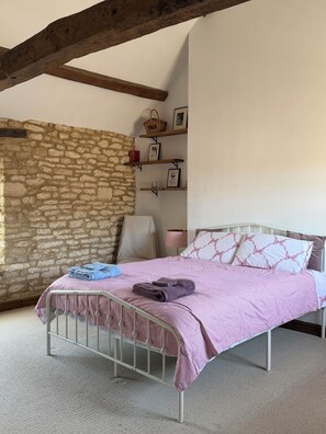 A bedroom with a stone wall, a metal bed frame, a pink checkered bedspread, and a wooden shelf with framed pictures.