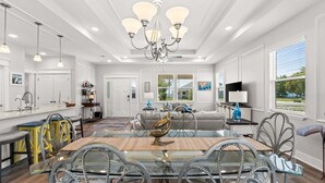 A spacious living room with a glass dining table, modern chandelier, and a large window with blinds. - Fall Deals + Snowbird Rates | Coastal Stay With Bay Views (Eastpoint)