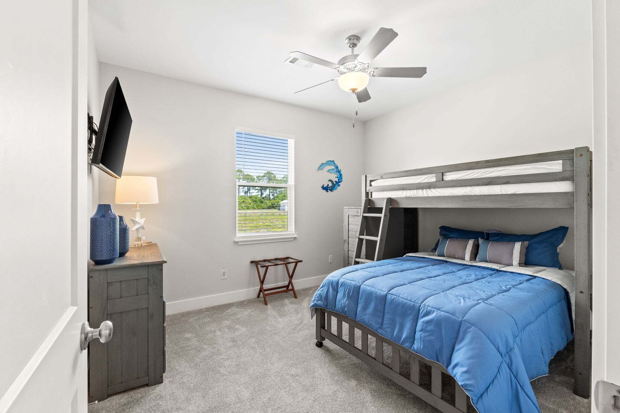 Une chambre avec un lit superposé, un ventilateur de plafond, un téléviseur fixé au mur, une fenêtre avec des stores et une petite table avec une lampe.