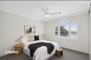 A bedroom with a bed, a ceiling fan, a window with blinds, a bedside table with a lamp, and a framed picture on the wall.