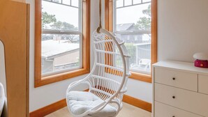 Een kamer met een witte rieten hangstoel, een witte dressoir met lades en grote ramen met uitzicht op een woonwijk.