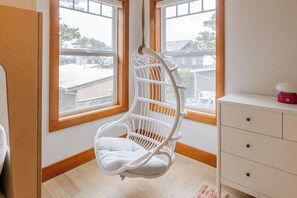 Ein Zimmer mit einem weißen Korbstuhl, der von der Decke hängt, einer weißen Kommode mit Schubladen und großen Fenstern mit Blick auf eine Wohngegend.