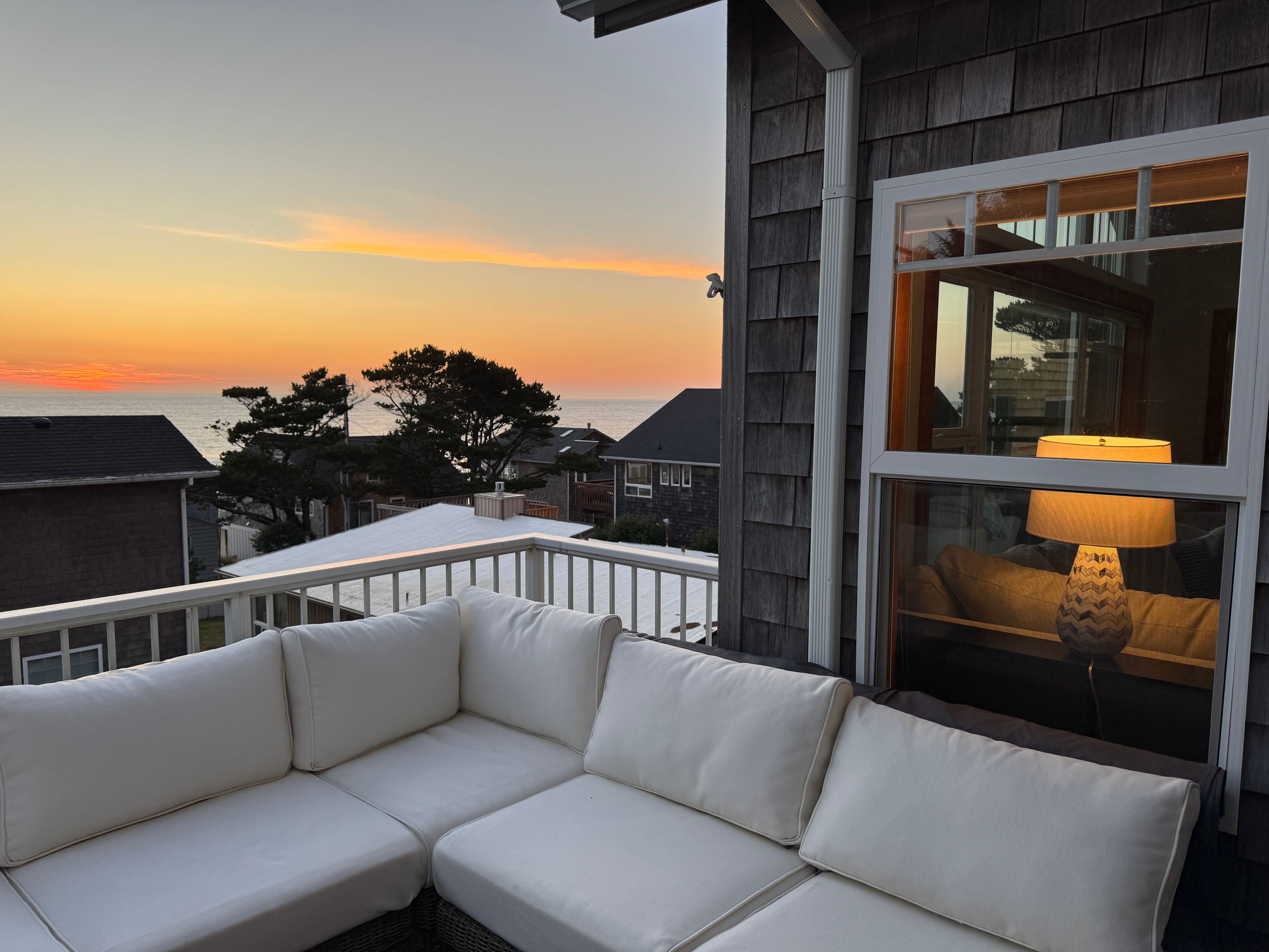 A balcony with a white cushioned sofa, a window with a lamp, and a view of the sunset over the ocean.