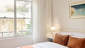 Una habitación de hotel con una cama, una mesita de noche con lámpara, un cuadro en la pared y una ventana con cortinas.