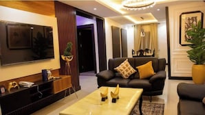 A modern living room with a flat-screen TV, a sofa with a decorative pillow, a coffee table with a plant, and a reception desk with a phone and a plant. - The Luxe Stay, 2 Bed Room Apartment (Lagos)