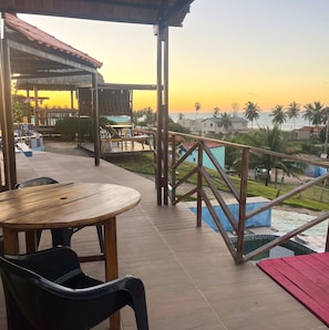 Una terraza con mesa y sillas de madera, una piscina y vistas al océano y palmeras.