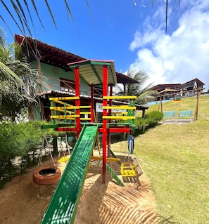 A playground with a slide and climbing structures, a building with a red roof, and a grassy area with a sign. - Pousada Bugaendrus Paradise (Touros)