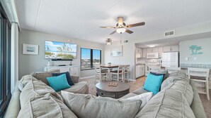 A living room with a sofa, coffee table, and a dining area with chairs. There is a kitchen with cabinets and appliances. - Island Winds East 311 | Walk to the Hang Out | Beachview | My Beach Getaways (Gulf Shores)