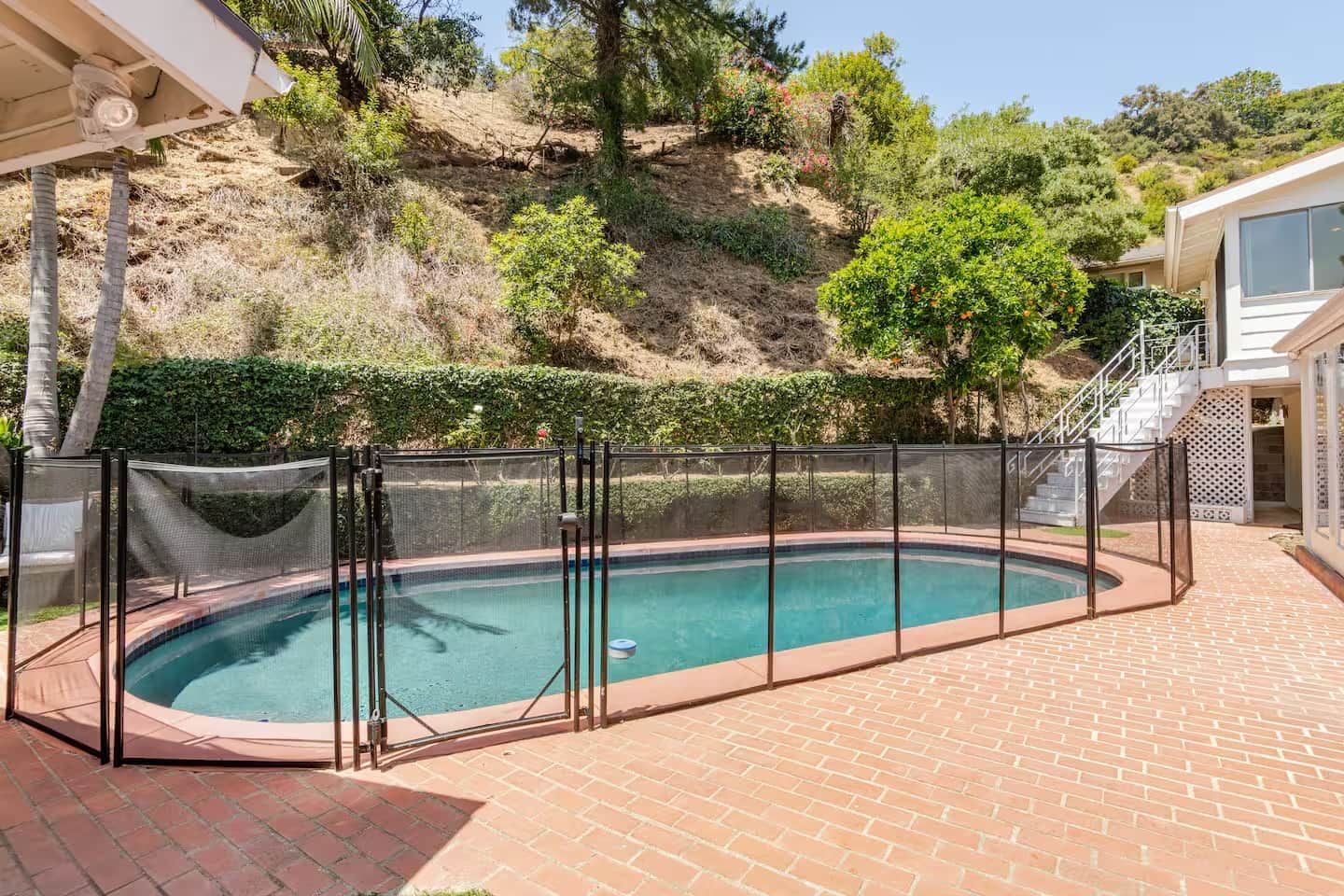 A swimming pool with a safety fence, surrounded by a brick patio, and a hillside with vegetation in the background.